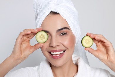 Photo of Woman in bathrobe with towel holding pieces of cucumber on light grey background. Spa treatment