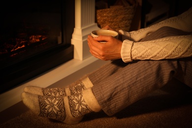 Photo of Woman in warm socks holding hot drink near fireplace with burning wood at home, closeup