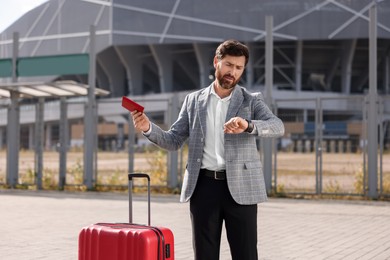 Photo of Being late. Worried businessman with red suitcase and passport looking at his watch outdoors