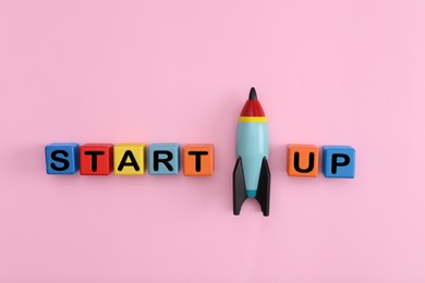 Photo of Colorful cubes with words Start Up and toy rocket on pink background, flat lay
