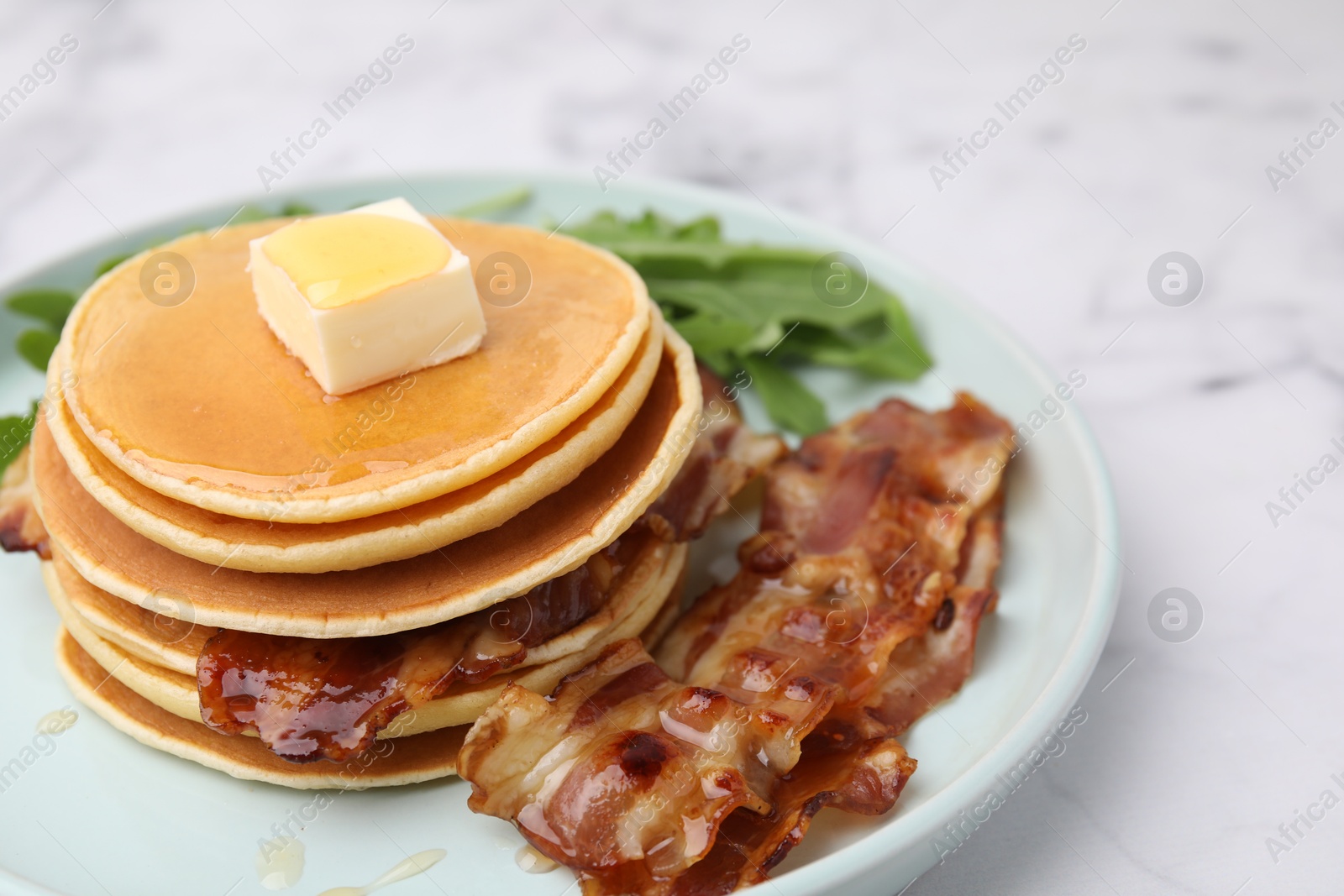 Photo of Tasty pancakes with butter, fried bacon and fresh arugula on white marble table, closeup