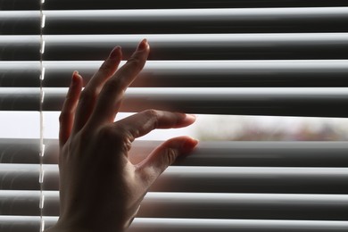 Photo of Woman separating slats of white blinds indoors, closeup