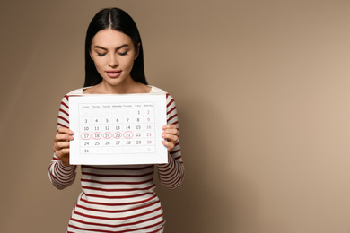 Young woman holding calendar with marked menstrual cycle days on beige background. Space for text