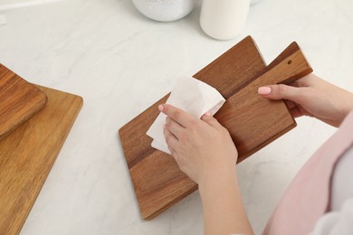 Photo of Woman wiping wooden cutting board with paper napkin at white table, above view