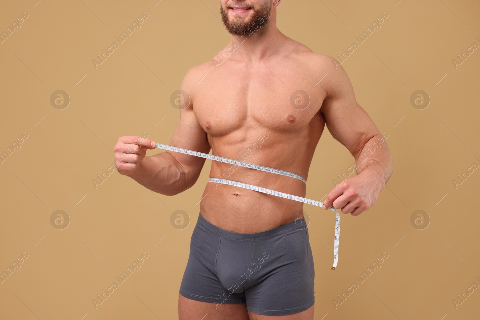 Photo of Smiling athletic man measuring waist with tape on brown background, closeup. Weight loss concept