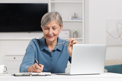 Beautiful senior woman taking notes near laptop at white table indoors