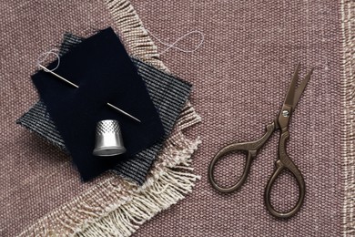 Photo of Silver thimble, scissors and needle on light brown cloth, flat lay. Sewing accessories