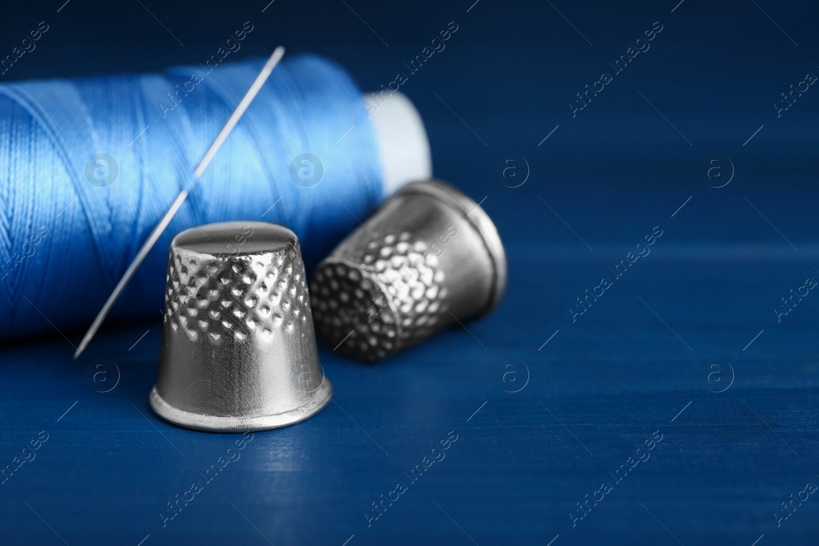Photo of Silver thimbles, sewing thread and needle on blue wooden table, closeup. Space for text