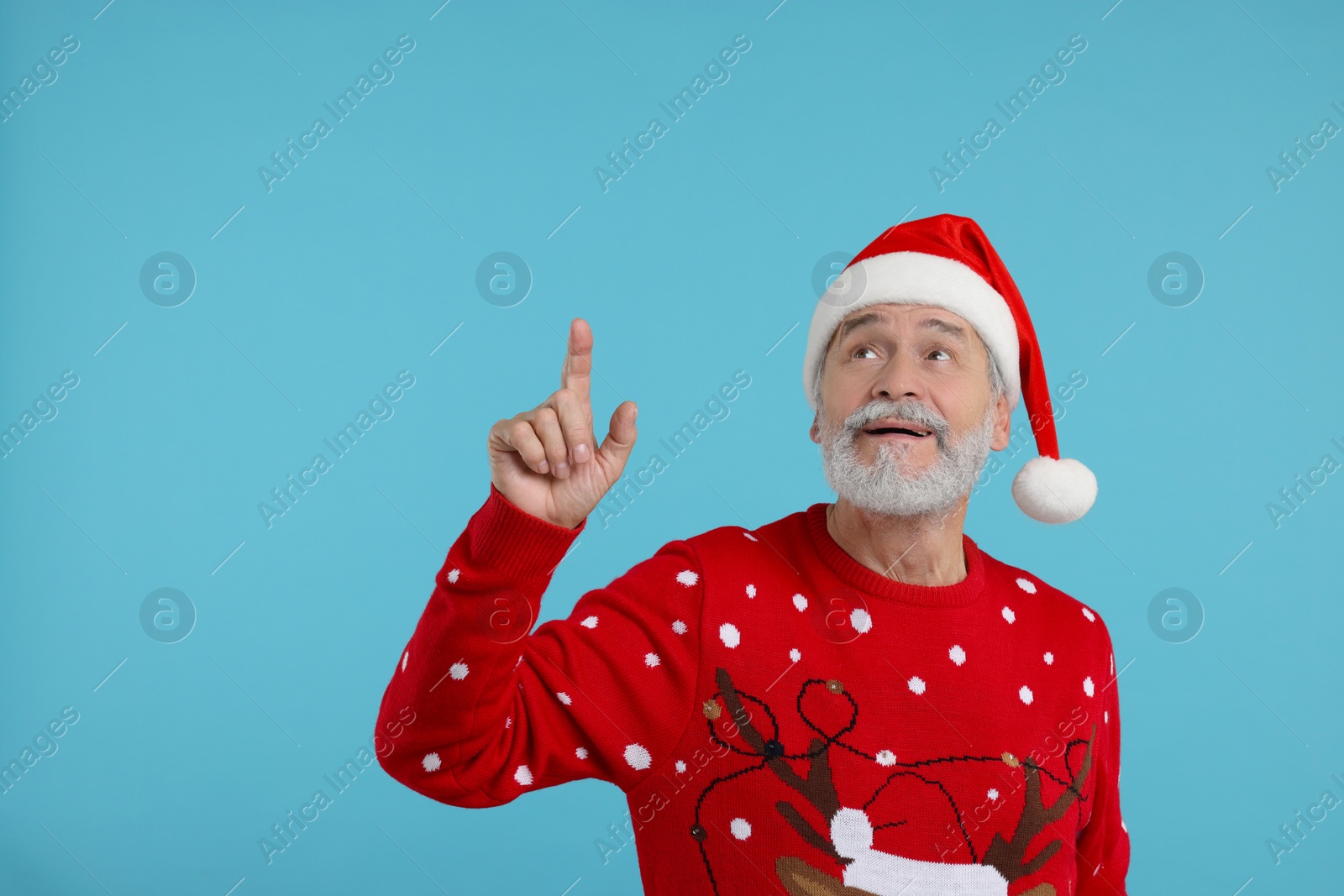Photo of Senior man in Christmas sweater and Santa hat pointing at something on light blue background. Space for text