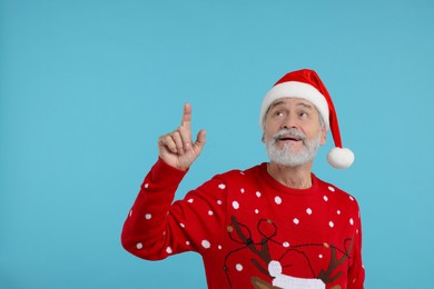 Senior man in Christmas sweater and Santa hat pointing at something on light blue background. Space for text