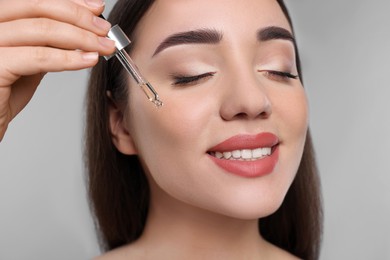 Happy young woman applying essential oil onto face on light grey background, closeup