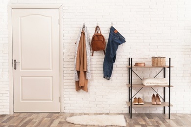 Photo of Stylish hallway interior with door, comfortable furniture and clothes on brick wall