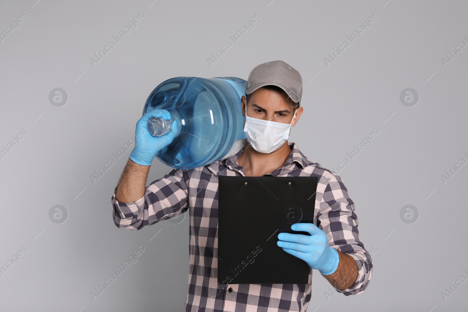 Photo of Courier in medical mask with bottle for water cooler and clipboard on light grey background. Delivery during coronavirus quarantine