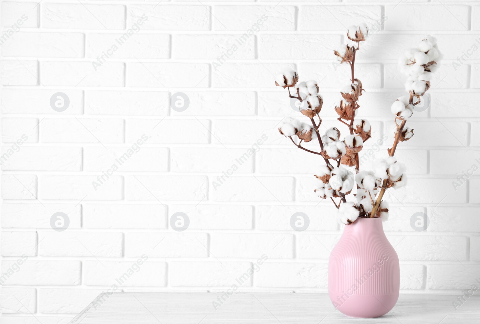 Photo of Beautiful cotton flowers in vase on wooden table against white brick wall, space for text