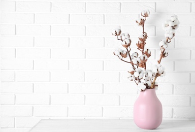 Beautiful cotton flowers in vase on wooden table against white brick wall, space for text