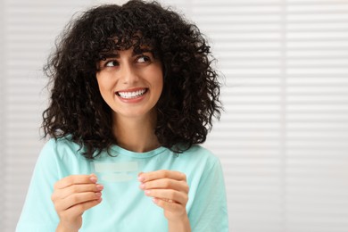 Young woman holding teeth whitening strips indoors, space for text