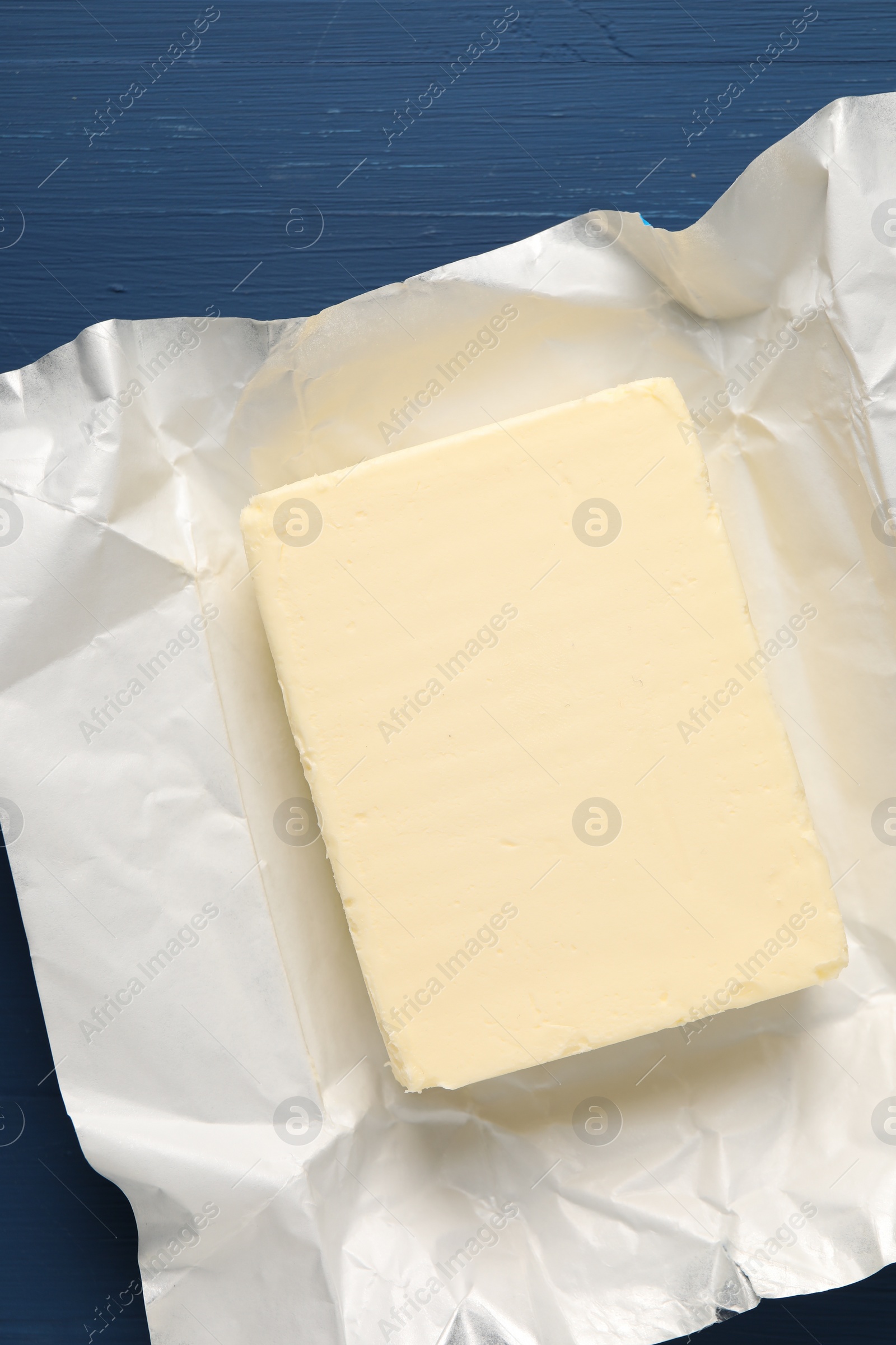 Photo of Block of tasty butter in open foil packaging on blue wooden table, top view