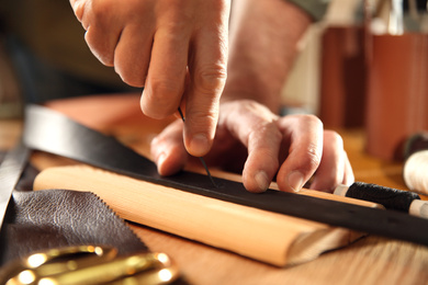 Photo of Man making holes in leather belt with stitching awl at table, closeup