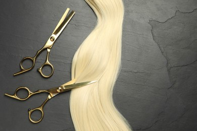 Professional hairdresser scissors with blonde hair strand on dark grey table, flat lay. Space for text