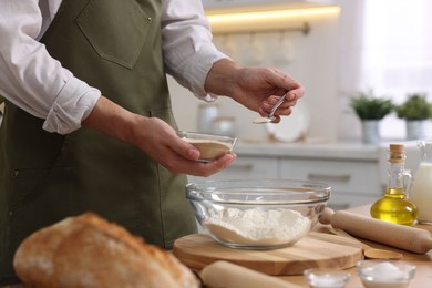 Photo of Making bread. Man putting dry yeast into bowl with flour at wooden table in kitchen, closeup
