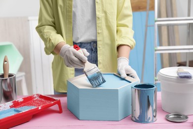 Photo of Woman painting honeycomb shaped shelf with brush at pink wooden table indoors, closeup
