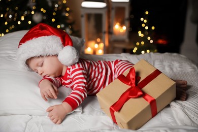 Photo of Baby in Christmas pajamas and Santa hat sleeping near gift box on bed indoors