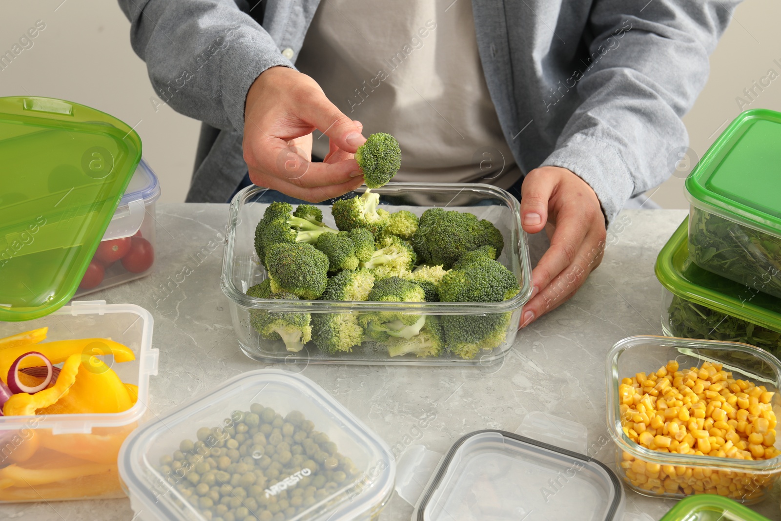 Photo of Man putting fresh broccoli into glass container at light grey table, closeup. Food storage