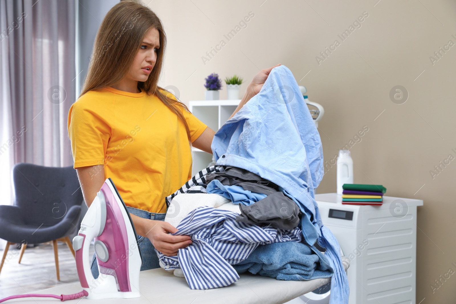 Photo of Emotional woman near board with iron and pile of clothes at home