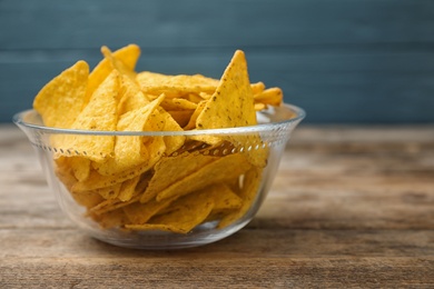 Glass bowl with tasty Mexican nachos chips on wooden table against blue background, space for text