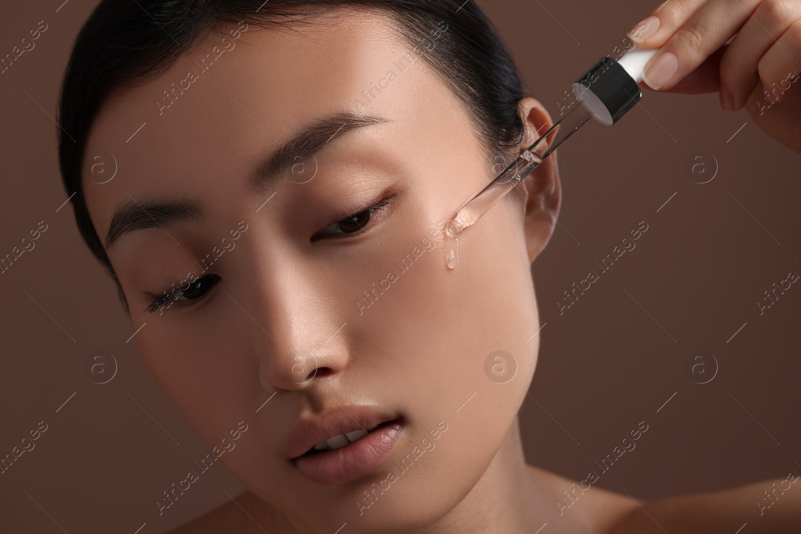 Photo of Beautiful young woman applying cosmetic serum onto her face on brown background, closeup