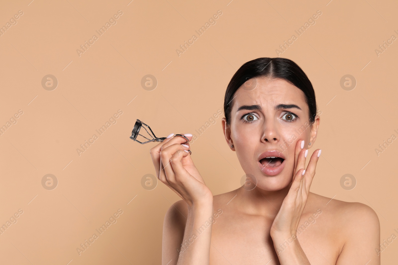 Photo of Emotional young woman with eyelash curler on beige background, space for text