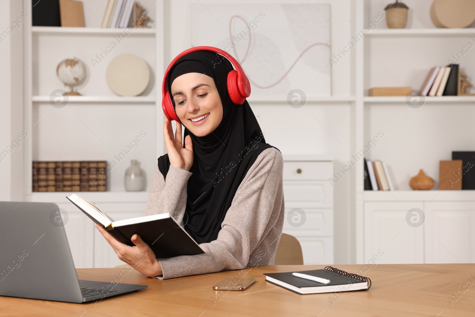 Photo of Muslim woman in headphones studying near laptop at wooden table in room