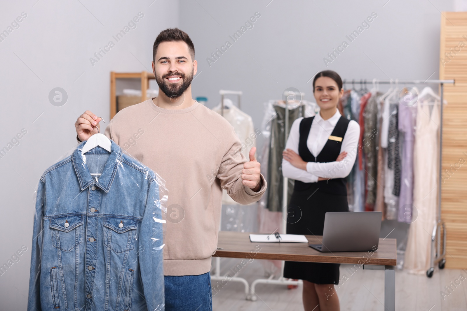 Photo of Dry-cleaning service. Man holding hanger with denim jacket and showing thumb up indoors. Happy worker at workplace