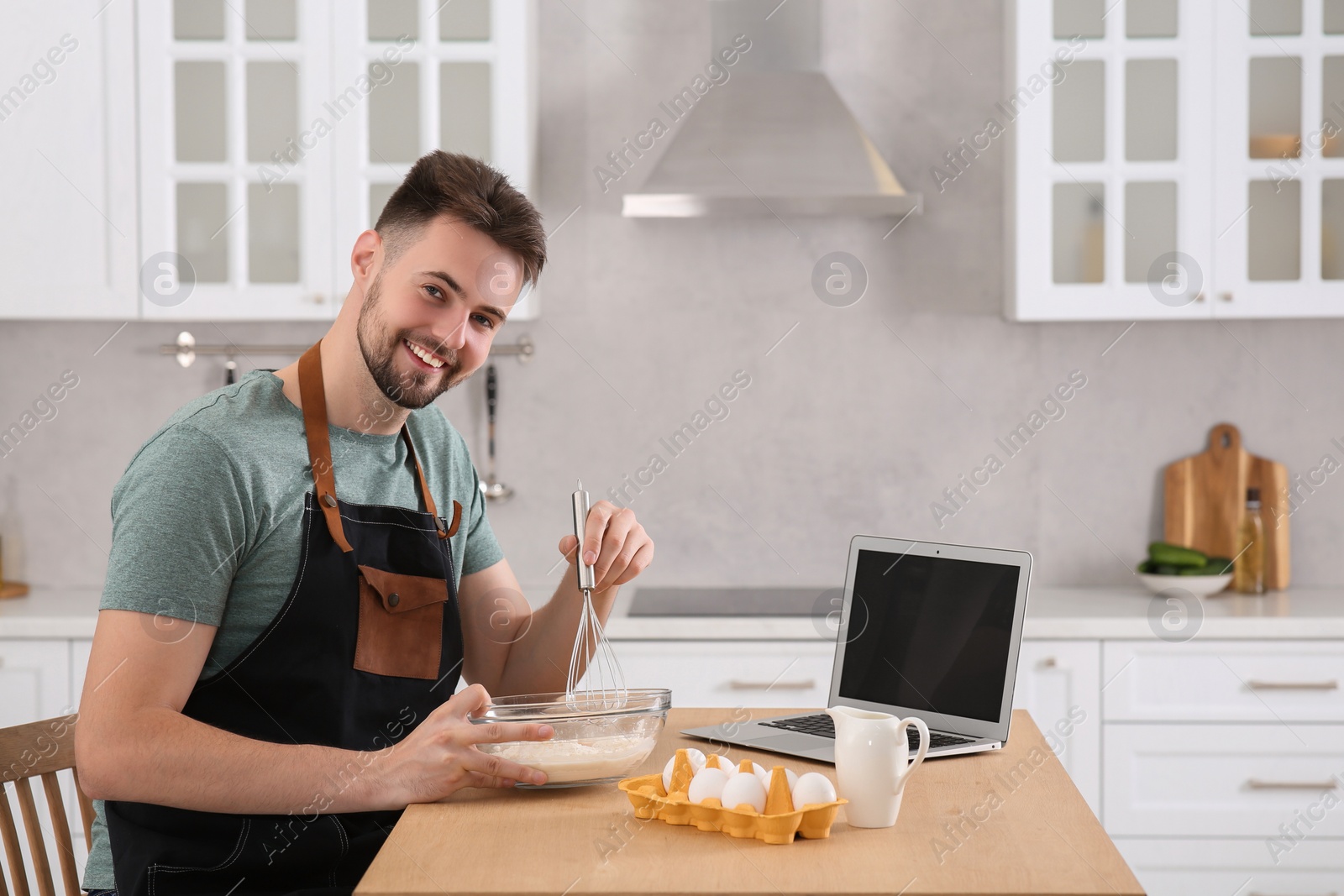 Photo of Happy man learning to cook with online video on laptop at table in kitchen. Time for hobby