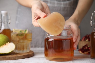 Photo of Woman putting Scoby fungus into jar with kombucha at white wooden table, closeup