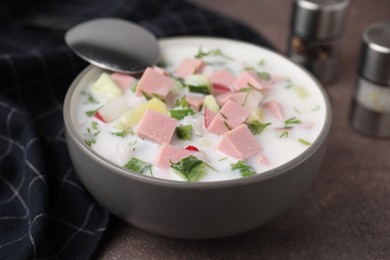 Photo of Delicious cold summer soup (okroshka) with boiled sausage in bowl and spoon on brown table, closeup