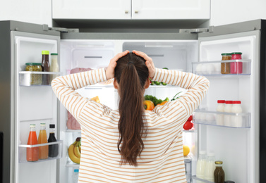 Worried young woman near open refrigerator in kitchen, back view