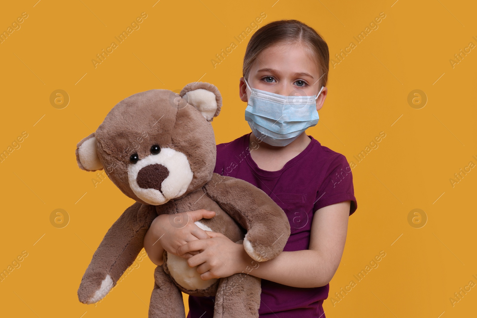 Photo of Cute girl with medical mask and teddy bear on orange background