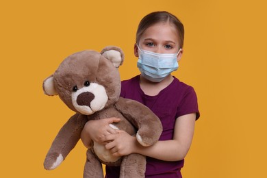 Photo of Cute girl with medical mask and teddy bear on orange background