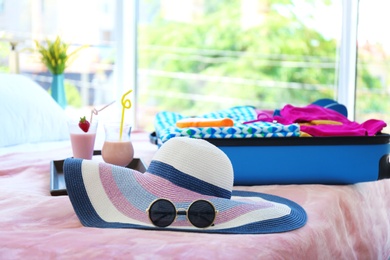 Hat with sunglasses, cocktails and open suitcase on bed indoors