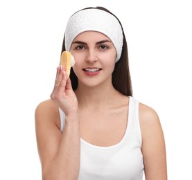 Young woman with headband washing her face using sponge on white background