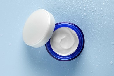 Moisturizing cream in open jar on light blue background with water drops, top view