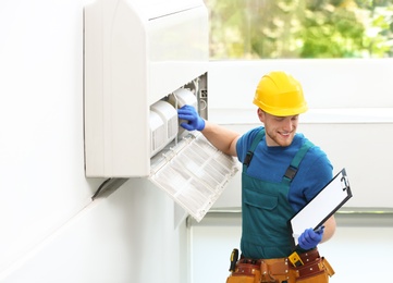 Photo of Professional technician with clipboard maintaining modern air conditioner indoors