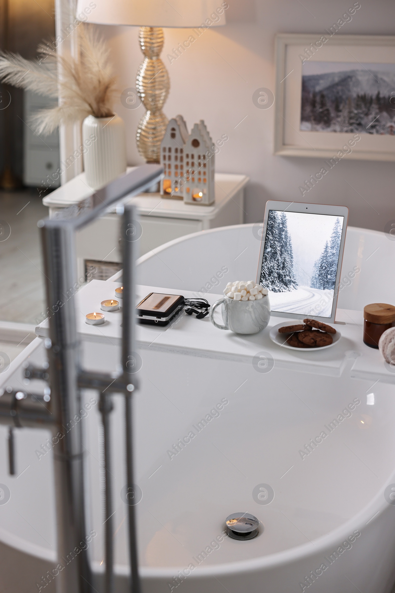 Photo of White wooden tray with tablet, cassette player and burning candles on bathtub in bathroom