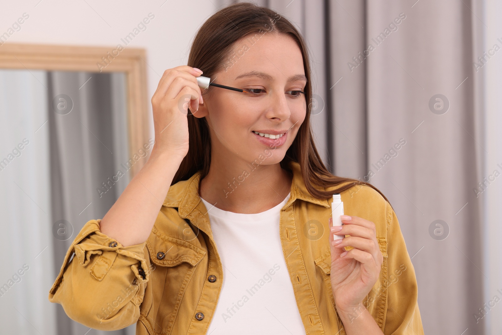 Photo of Beautiful woman applying serum onto eyelashes indoors