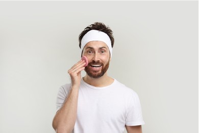 Man with headband washing his face using sponge on light grey background