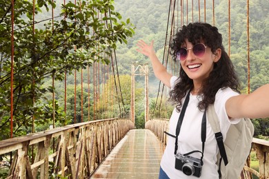 Beautiful woman in sunglasses with camera taking selfie on rusty metal bridge over river in mountains