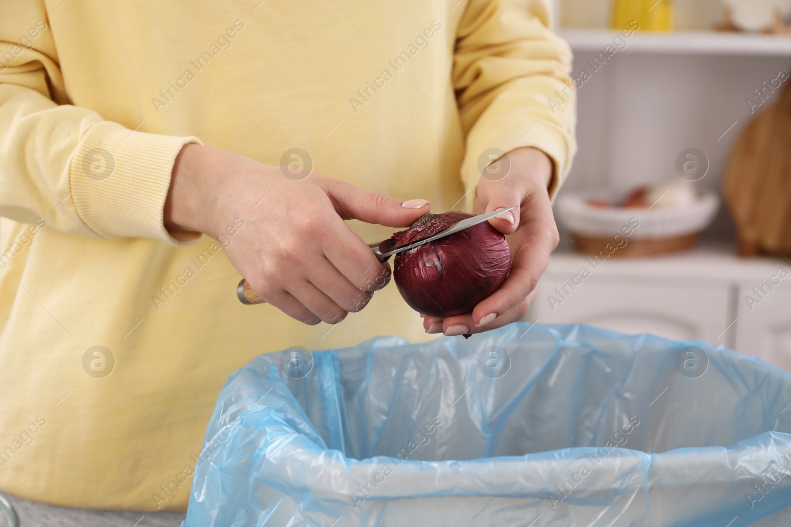 Photo of Woman peeling fresh onion above garbage bin indoors, closeup