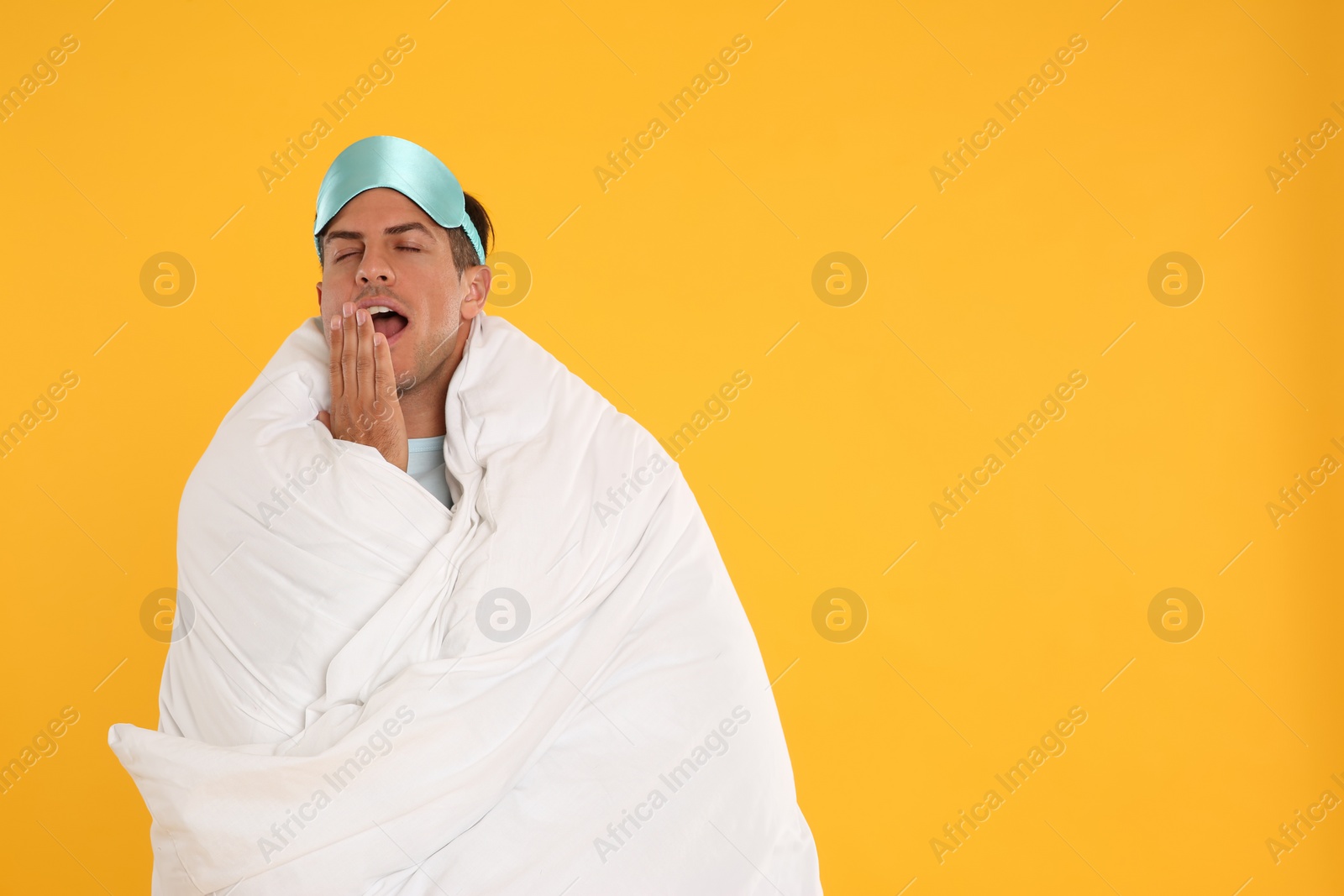 Photo of Man wrapped with blanket in sleeping mask yawning on yellow background. Space for text