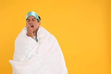 Man wrapped with blanket in sleeping mask yawning on yellow background. Space for text